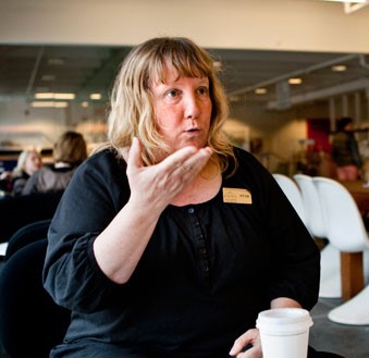 Tolken Lena Ekevid teckenspråkar med någon på ett café på Stockholms universitet. Hon har axellångt utsläppt mörkblont hår med lugg, svart tröja och sin tolkskylt på sig. På bordet framför Lena står en pappersmugg med lock.
